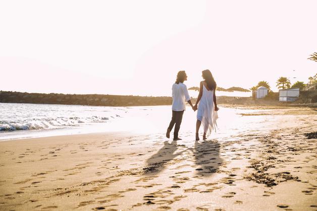 Un couple marche main dans la main sur la plage, profitant d'une babymoon grossesse romantique au coucher du soleil.
