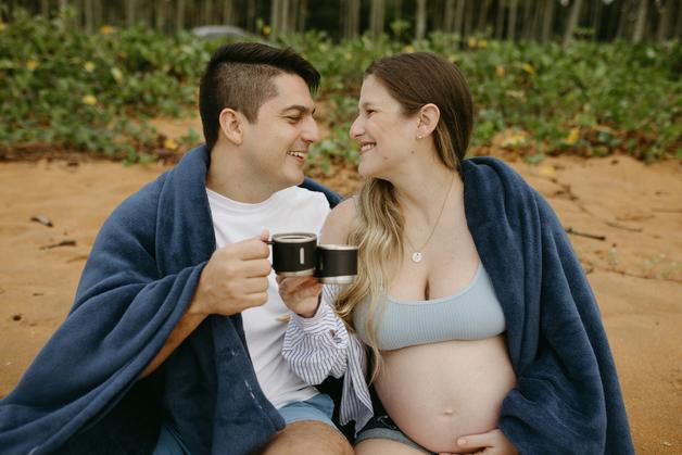 Un couple souriant partage un café grossesse sur la plage, enveloppé dans des couvertures confortables.