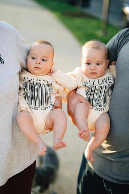 Deux bébés portant des bodys identiques, représentant des faux jumeaux, sont tenus par leurs parents.
