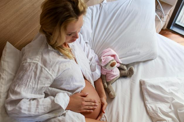 Femme enceinte assise sur un lit, touchant son ventre pour reconnaître contractions. Peluche à ses côtés.