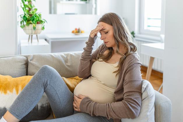 Femme enceinte assise sur un canapé, ressentant des symptômes de mononucléose enceinte, avec une main sur le front.